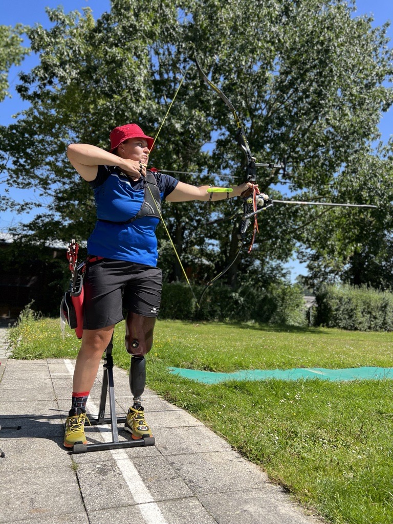 Eine Person mit einer Beinprothese steht auf einem sonnigen Bogenschießplatz, spannt einen Bogen und zielt auf ein Ziel. Sie trägt eine rote Mütze, ein blaues Hemd und schwarze Shorts. Im Hintergrund ist üppiges Grün zu sehen.