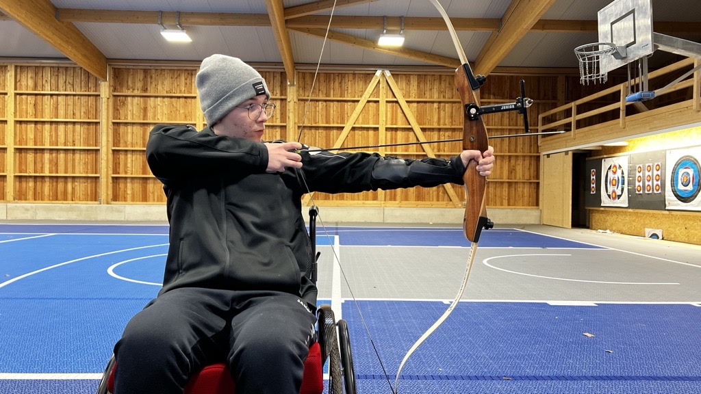 Person im Rollstuhl übt Bogenschießen in einer Indoor-Sportanlage und zielt mit einem Bogen auf ein Ziel.