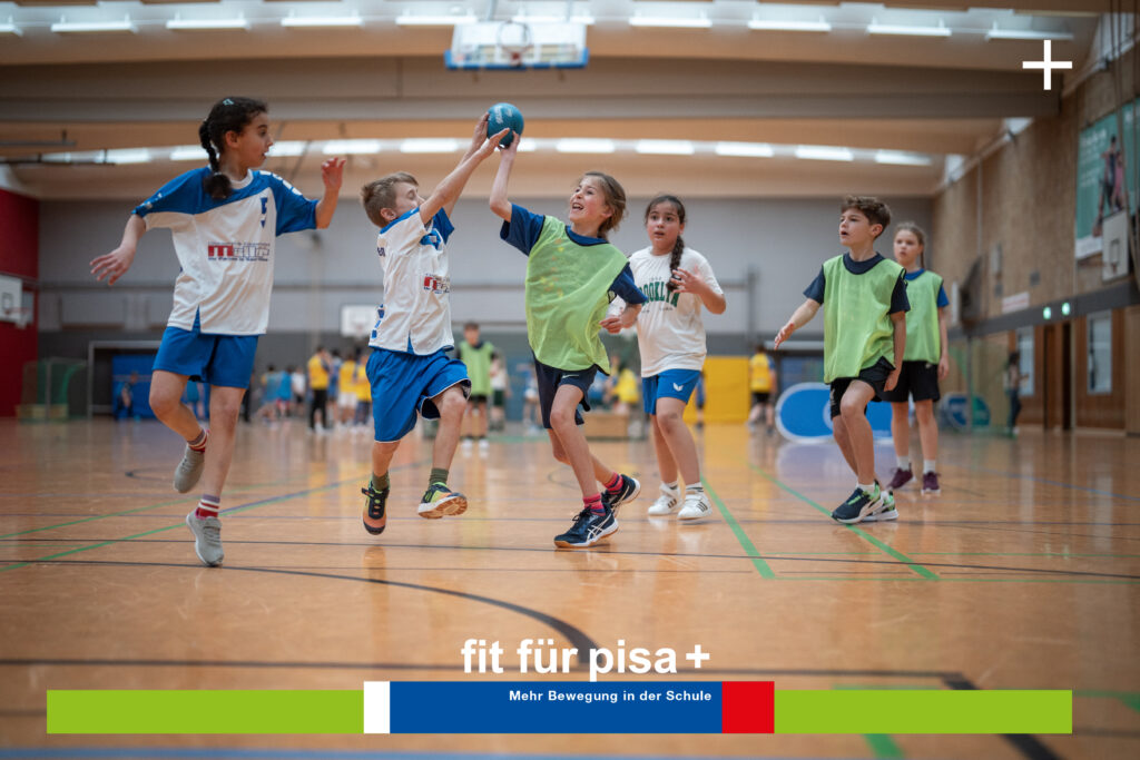 Kinder spielen in einer Turnhalle Handball. Sie tragen blaue und grüne Leibchen. Im Mittelpunkt steht ein Spieler, der hochspringt, um den Ball abzufangen. Unter Bannern wird für eine Fitnessinitiative geworben und die Bedeutung der Ernährung hervorgehoben.