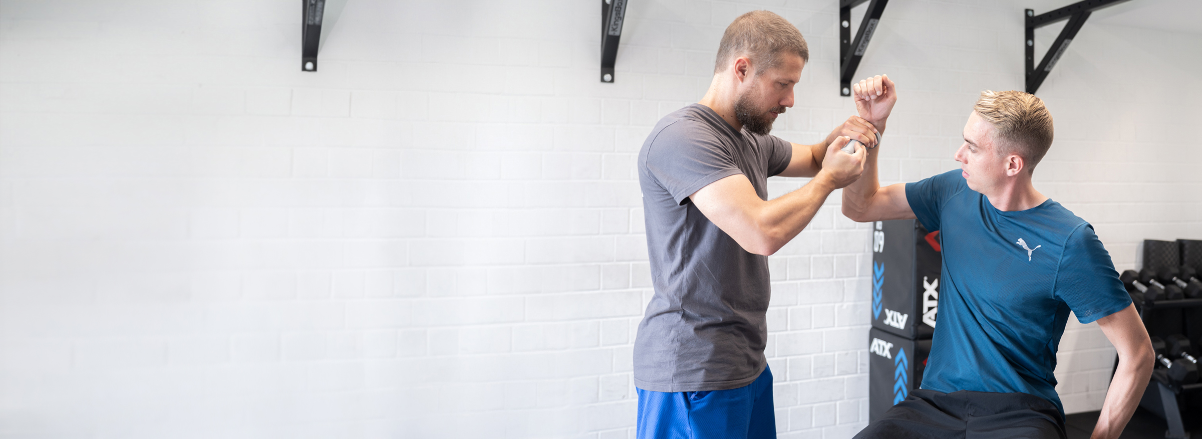 Two men practicing self-defense techniques in a gym with white walls and exercise equipment in the background.