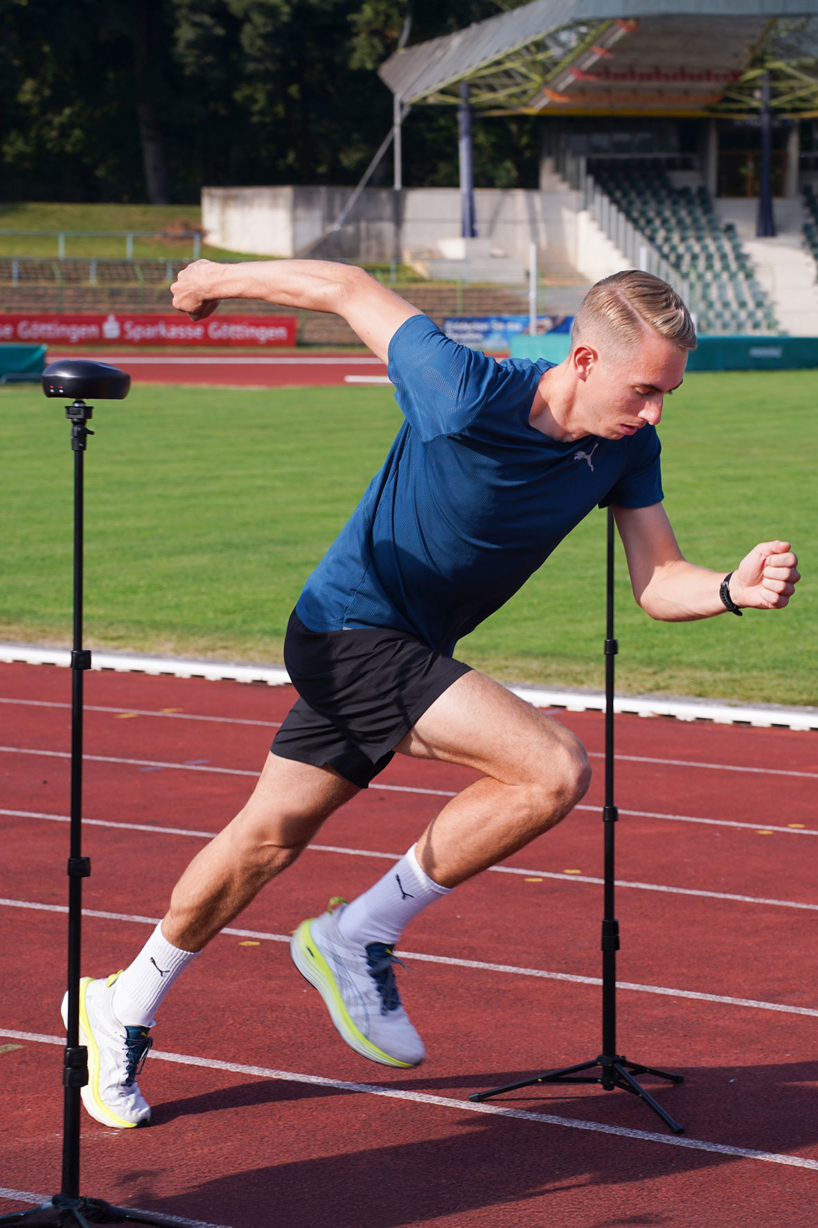 Ein Mann in Sportkleidung sprintet auf einer Bahn zwischen zwei vertikalen Sensoren hindurch; im Hintergrund sind leere Stadionsitze und ein Feld zu sehen.