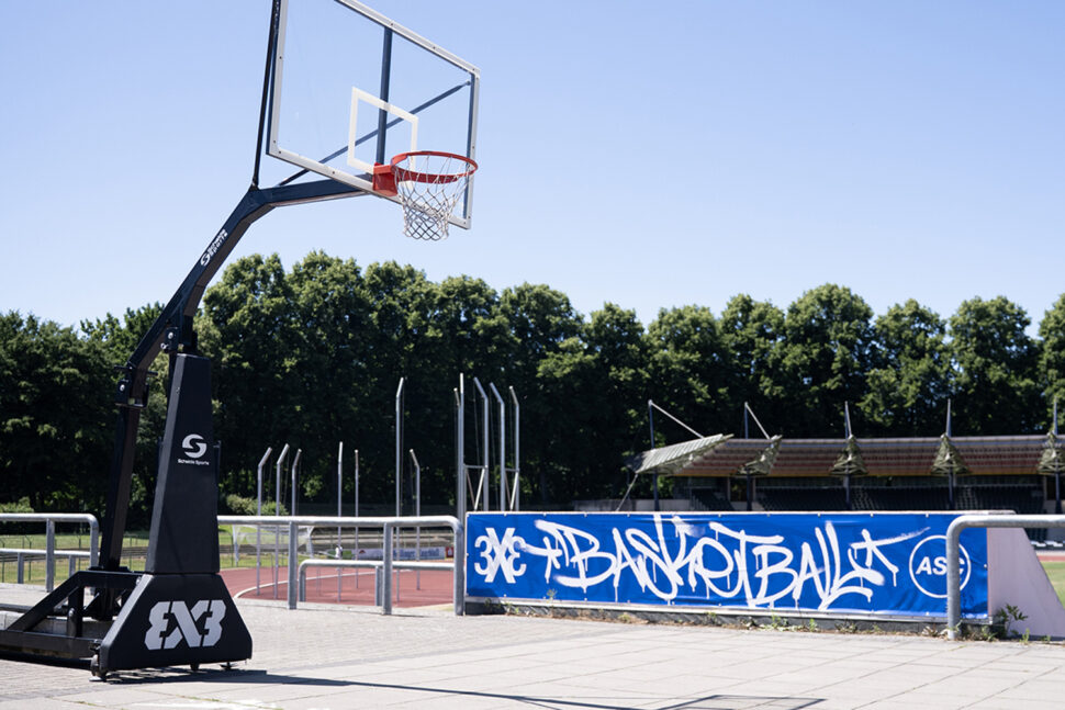 Ein Basketballkorb steht auf einem gepflasterten Platz neben einem blauen Banner mit der Aufschrift 