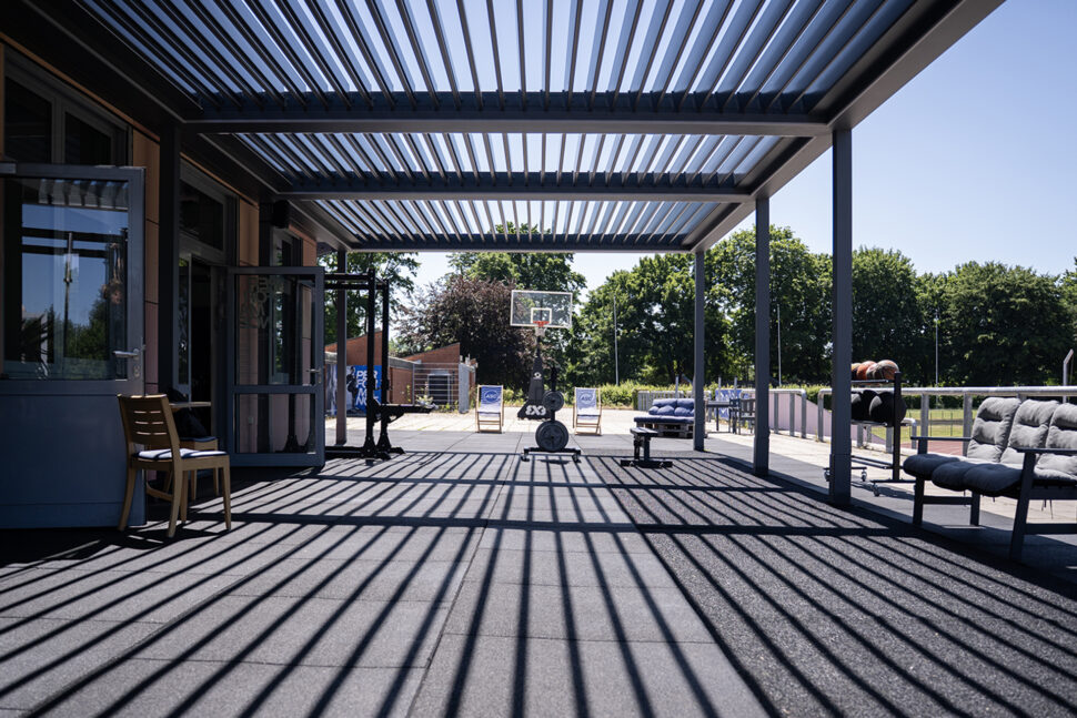 Fitnessbereich im Freien mit leistungsorientierten Trainingsgeräten unter einer Pergola, die gestreifte Schatten auf dem Boden erzeugt; im Hintergrund sind ein Basketballkorb und Bäume zu sehen.