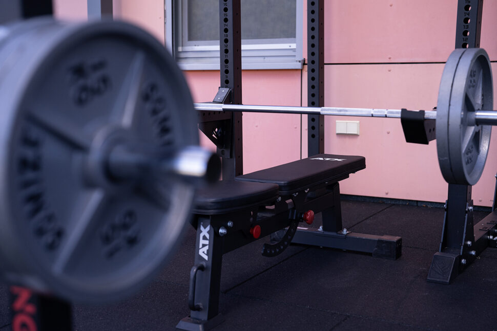 Ein Bankdrücken mit einer belasteten Langhantel in einem Fitnessstudio, das auf optimale Leistung ausgelegt ist, mit einer rosa Wand und einem Fenster im Hintergrund.