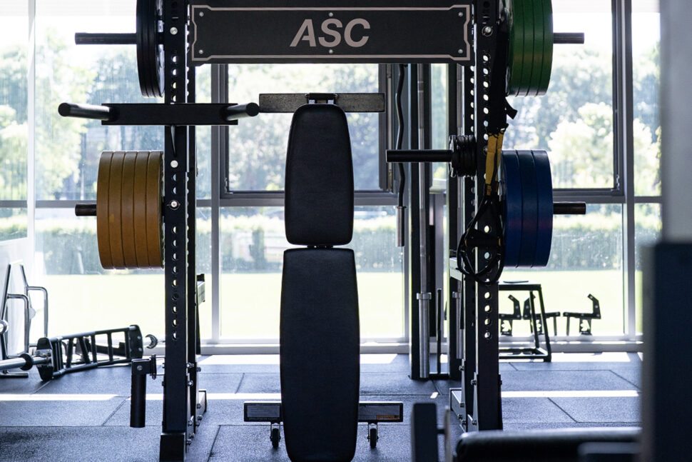 Eine Hantelbank und ein Squat Rack mit verschiedenfarbigen Hantelscheiben in einem auf Höchstleistung ausgelegten Fitnessstudio mit großen Fenstern, die den Blick ins Grüne freigeben.