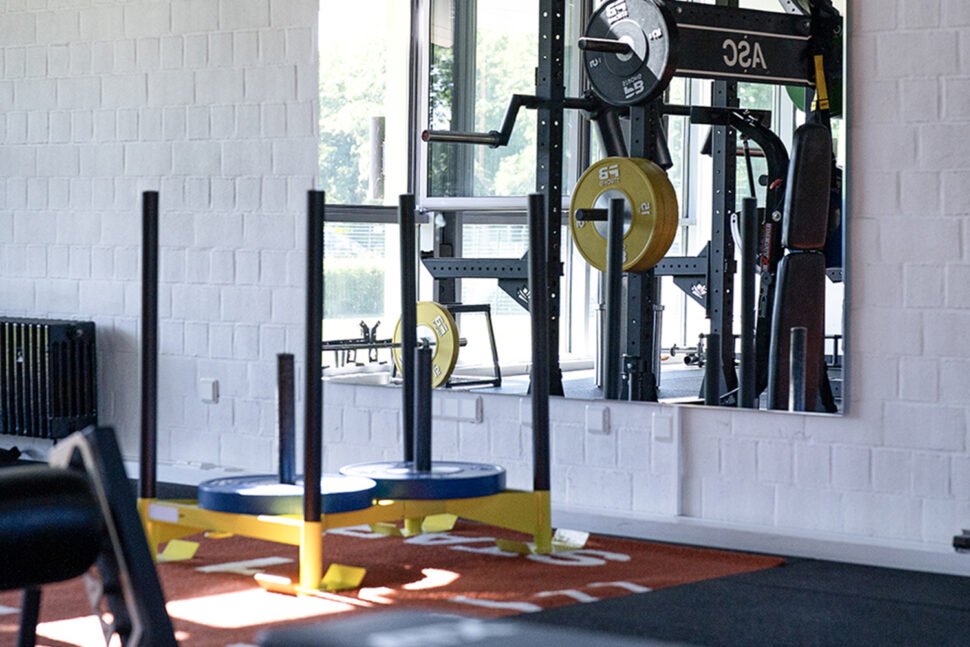 Ein Fitnessstudio, das auf Leistung ausgelegt ist, mit Geräten zum Gewichtheben wie einem Power Rack mit gelben Platten und einem Schlitten mit blauen Platten auf einer roten Bahn.
