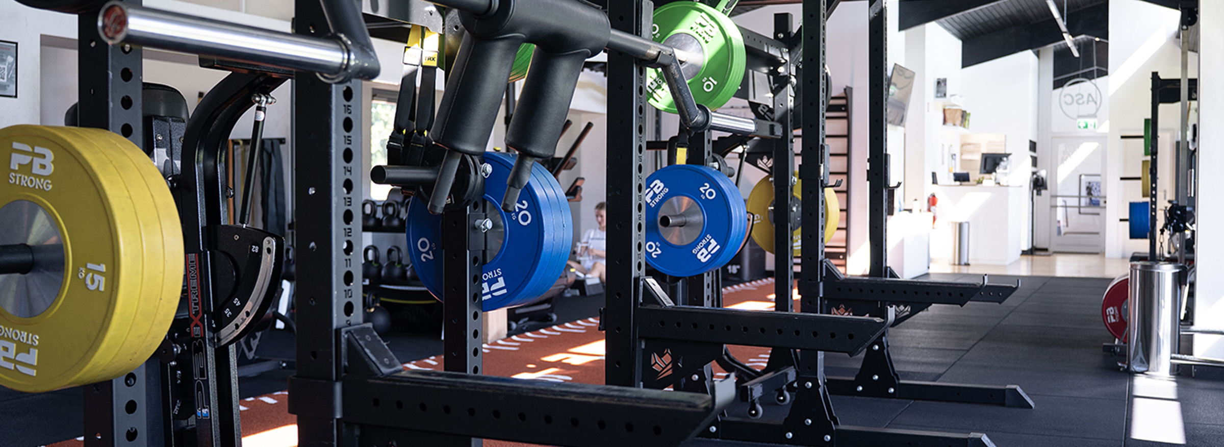 Ein Fitnessraum mit Hantelständern, bunten Hantelscheiben und Trainingsgeräten auf einem Gummiboden, der die Leistung steigern soll, mit natürlichem Licht, das durch große Fenster einfällt.