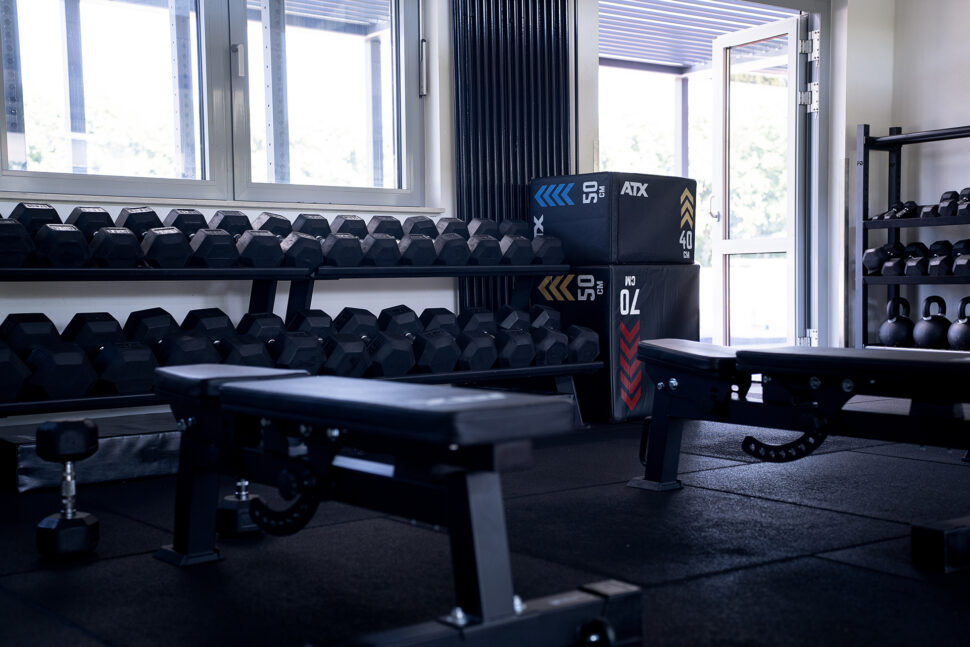 Ein leistungsorientiertes Fitnessstudio mit schwarzen Hanteln auf Gestellen, verstellbaren Bänken, plyometrischen Boxen, Kettlebells und natürlichem Licht, das durch Fenster und eine offene Tür einfällt.