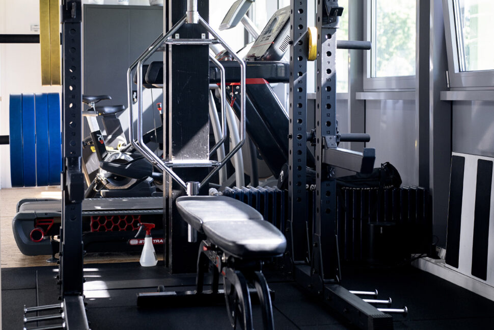 Der Innenraum des Fitnessstudios ist auf Höchstleistung ausgelegt und verfügt über verschiedene Trainingsgeräte, darunter eine Bank, ein Power Rack, eine Hex-Bar, ein Laufband und Hantelscheiben in der Nähe großer Fenster.