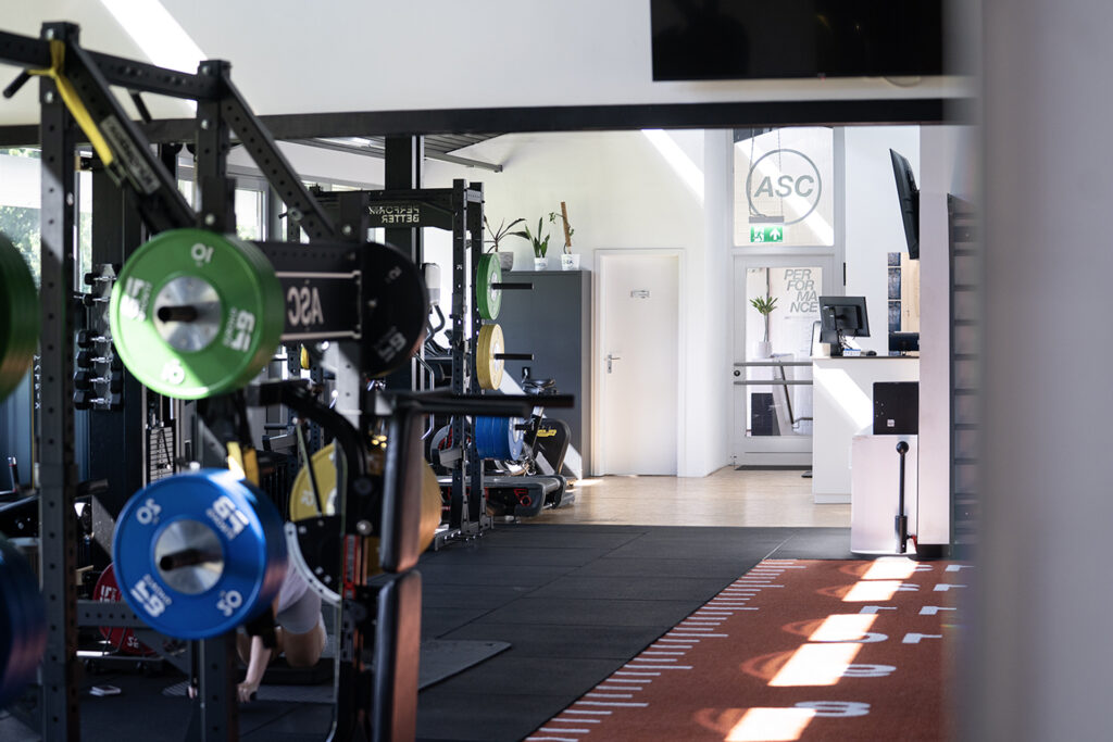 Modernes Fitnessstudio mit leistungsorientierten Kraftdreikampfgeräten, bunten Platten und einer Laufbahn; im Hintergrund ist der Empfangsbereich zu sehen.