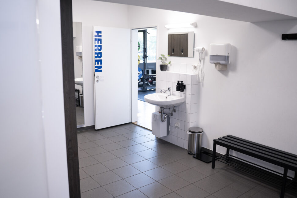 Eine saubere Herrentoilette mit Waschbecken, Spiegel, Seifenspender, Händetrockner, Abfalleimer, schwarzer Sitzbank und einer Tür mit der Aufschrift HERREN