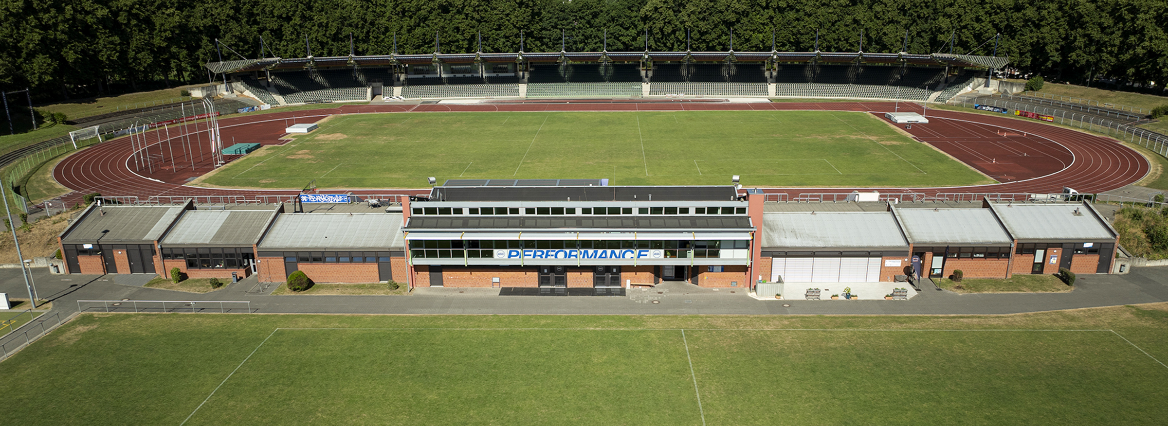 Luftaufnahme eines Leichtathletikstadions im Freien mit einer Laufbahn, einem Rasenplatz, Tribünen und einem zentralen Gebäude mit der Aufschrift 