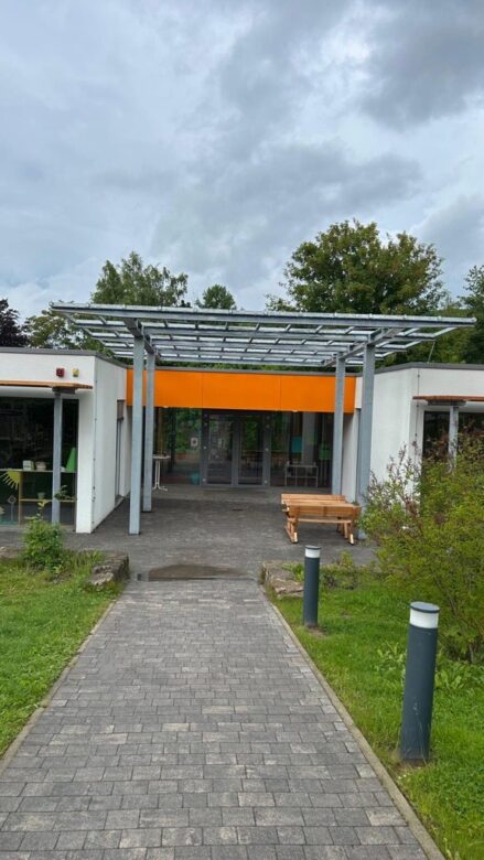Ein gepflasterter Weg führt zum Eingang eines Gebäudes mit einem Vordach aus Metall und Glas. Das Gebäude hat weiße Wände und einen orangefarbenen Akzent über der Tür. In der Nähe befinden sich Bänke und Grünflächen.