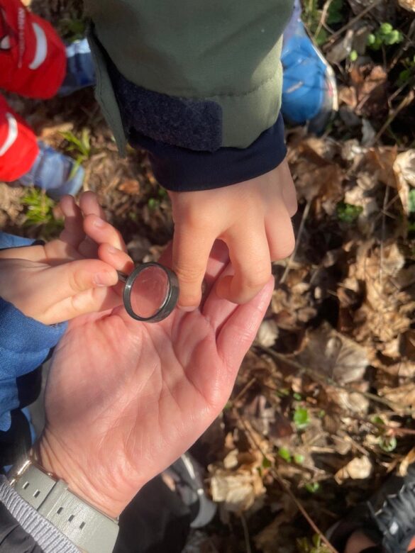 Eine erwachsene Hand hält eine Lupe, während ein Kind danach greift, um sie zu berühren; umgeben von Laub und anderen Kindern in der Nähe.