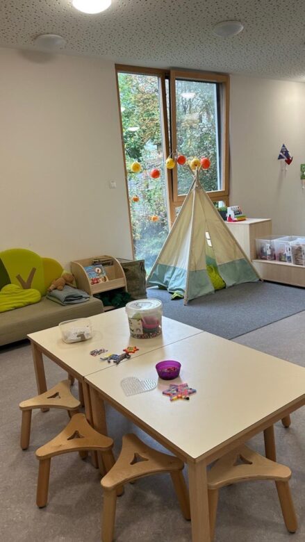 Ein Vorschulraum mit einem Tisch, Hockern, Bastelmaterial, einem grünen Sofa, einem kleinen Zelt für drinnen und großen Fenstern mit hängenden Dekorationen.