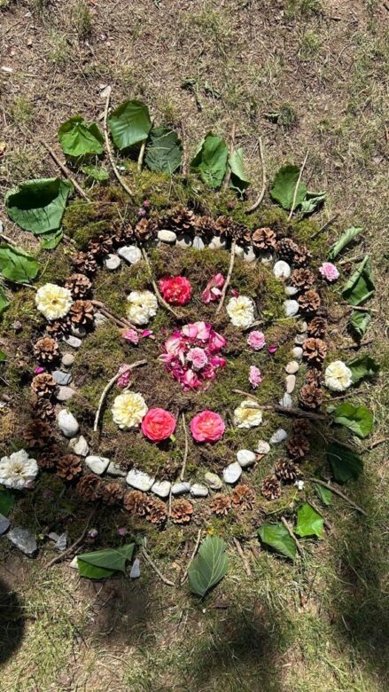 Ein kreisförmiges Mandala auf Gras, bestehend aus Blättern, Blumen, Tannenzapfen, Zweigen, Moos und Steinen, die in konzentrischen Mustern angeordnet sind.