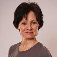 Eine Frau mittleren Alters mit kurzen schwarzen Haaren, die ein graues Fitness-T-Shirt trägt, steht vor einem schlichten hellen Hintergrund, schaut in die Kamera und lächelt leicht.