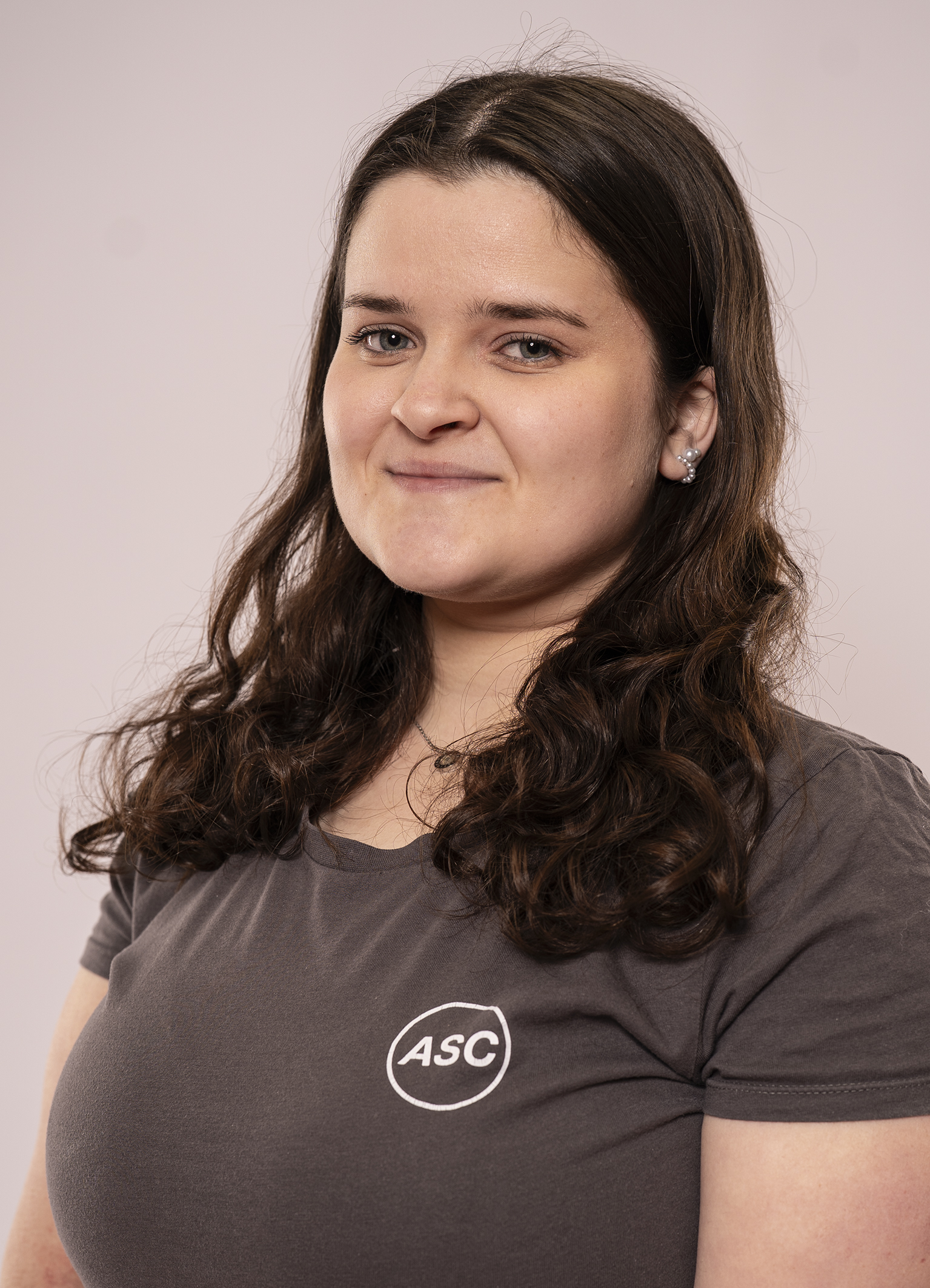 Eine Frau mit langen braunen Haaren, die ein graues ASC-Fitness-T-Shirt trägt, steht vor einem schlichten hellen Hintergrund, schaut in die Kamera und lächelt leicht.