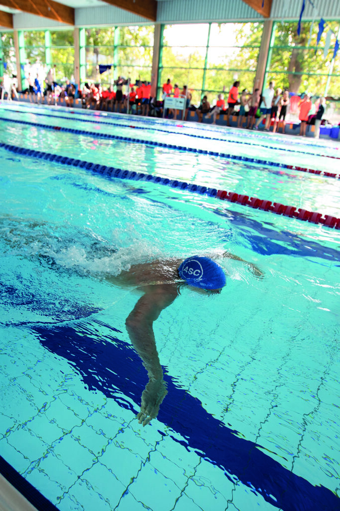 Ein Schwimmer mit einer blauen Kappe schwimmt während eines Wettkampfs in einem Hallenbad Freistil, während im Hintergrund Zuschauer und Offizielle zu sehen sind.