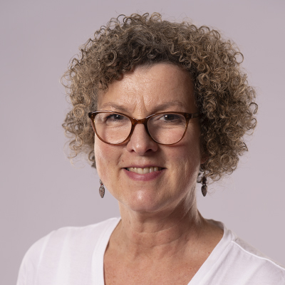 Eine Frau mit kurzen lockigen Haaren, Brille und Ohrringen lächelt in die Kamera. Sie trägt ein weißes Oberteil vor einem schlichten hellen Hintergrund und strahlt Wärme aus - ein sympathisches Mitglied des Teams.