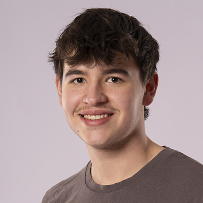 Ein junger Mann mit kurzem, dunklem, gewelltem Haar lächelt in die Kamera. Er trägt ein braunes T-Shirt, steht vor einem schlichten hellen Hintergrund und strahlt eine natürliche Fitness und jugendliche Energie aus.