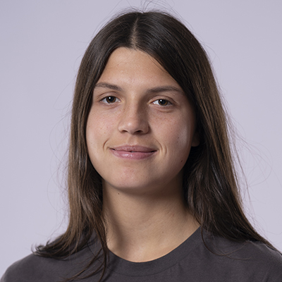 Eine junge Frau mit glattem braunem Haar und neutralem Gesichtsausdruck, die ein dunkles Fitness-T-Shirt trägt, posiert vor einem schlichten hellen Hintergrund.