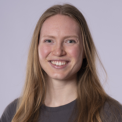 Eine Frau mit langen, glatten blonden Haaren und einem grauen Fitness-Shirt lächelt in die Kamera vor einem hellvioletten Hintergrund.