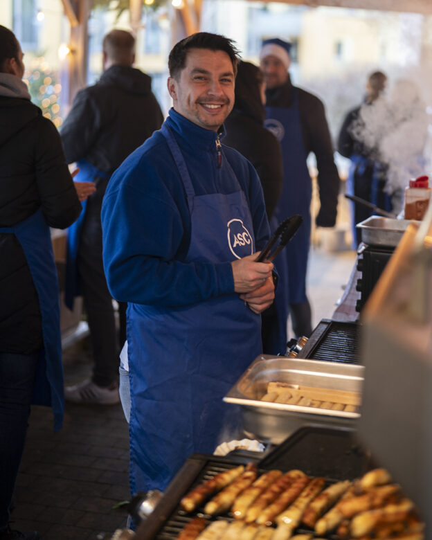 Ein Mann mit einer blauen Schürze und einer Zange in der Hand steht an einem Grill mit Würstchen und lächelt an einem Imbissstand im Freien. Im Hintergrund sind andere Personen und Kochgeräte zu sehen.