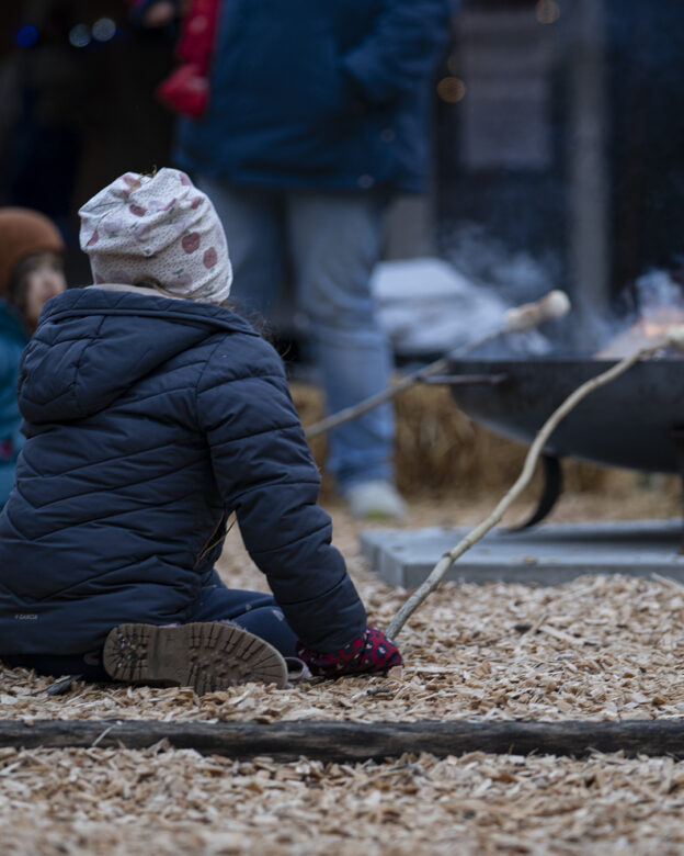 Ein Kind in Wintermantel und Mütze sitzt auf Holzspänen und röstet mit einem Stock Marshmallows über einer Feuerstelle.