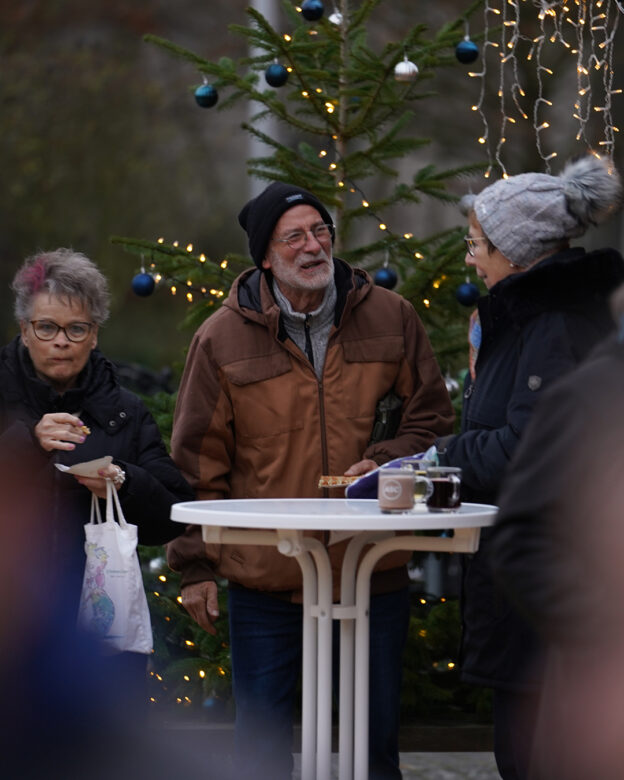 Drei ältere Erwachsene stehen um einen hohen Tisch im Freien, essen und unterhalten sich neben einem geschmückten Weihnachtsbaum mit Lichtern und Ornamenten.