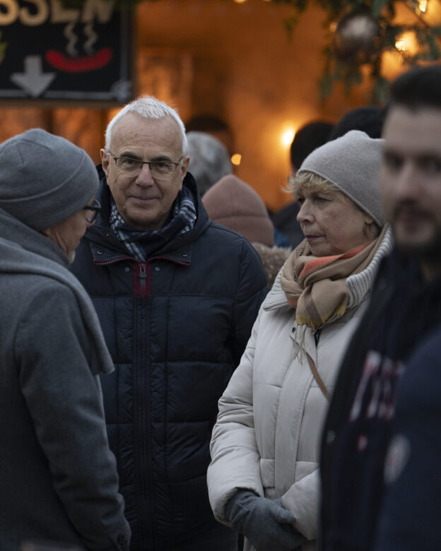 Drei Erwachsene in Winterkleidung stehen im Freien inmitten einer Menschenmenge, mit warmen Lichtern im Hintergrund und einem teilweise sichtbaren Schild über ihnen.