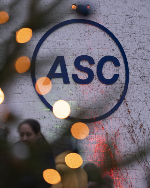 Ein rundes blaues ASC-Logo-Schild an einer weißen Backsteinmauer, mit verschwommenen Lichtern und Menschen, die teilweise im Vordergrund zu sehen sind.