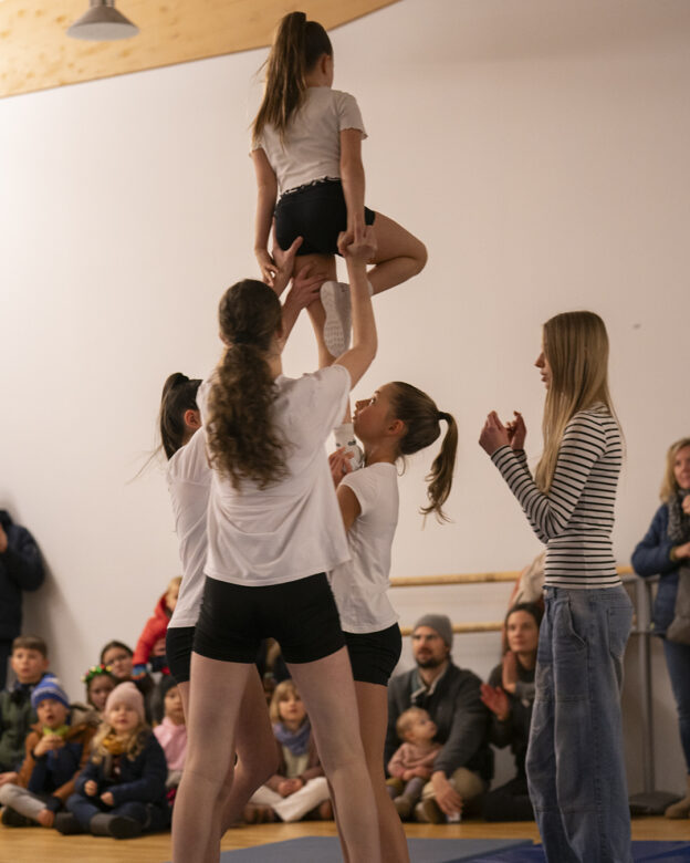 Vier junge Turnerinnen führen in einer Halle eine Hebeübung vor, während eine Lehrerin zuschaut und ein Publikum im Hintergrund sitzt.