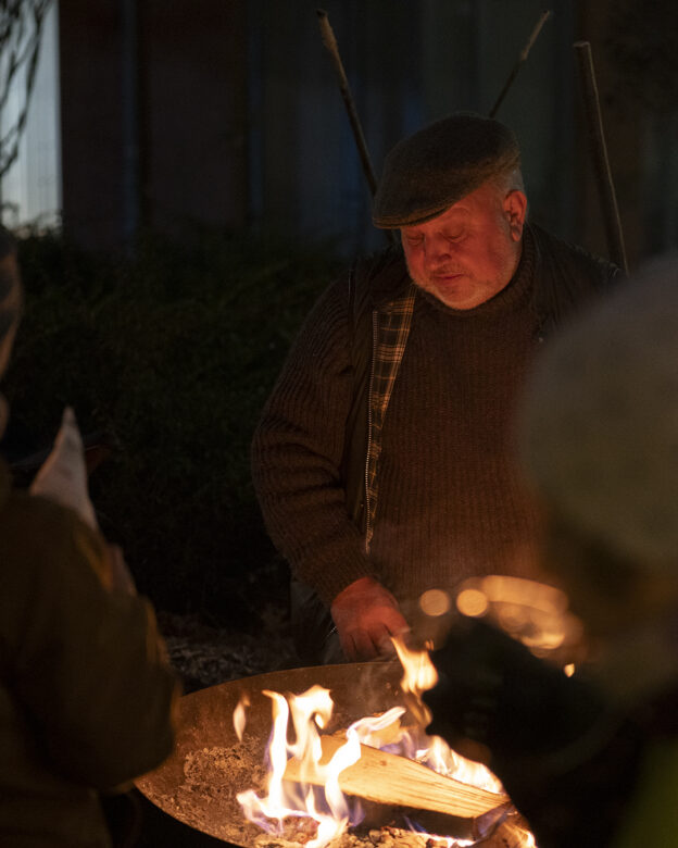 Ein älterer Mann mit Mütze und Pullover steht nachts an einer Feuerstelle im Freien, wo sich Menschen um die Flammen scharen.