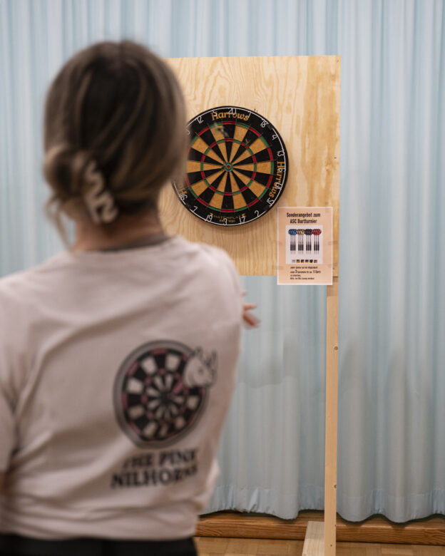 Eine Person steht in einem Raum vor einer auf einer Holzplatte montierten Dartscheibe und bereitet sich darauf vor, einen Dartpfeil zu werfen. Neben der Dartscheibe ist ein Schild mit den Regeln für Dartscheiben angebracht.