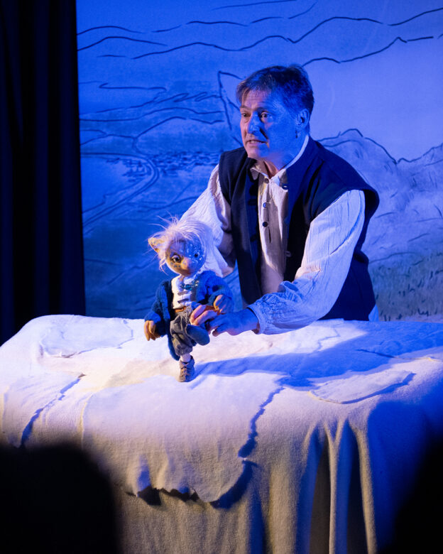 Ein Mann bedient eine kleine Marionette auf einem mit weißem Stoff bespannten Tisch mit einer blauen Berglandschaft im Hintergrund.