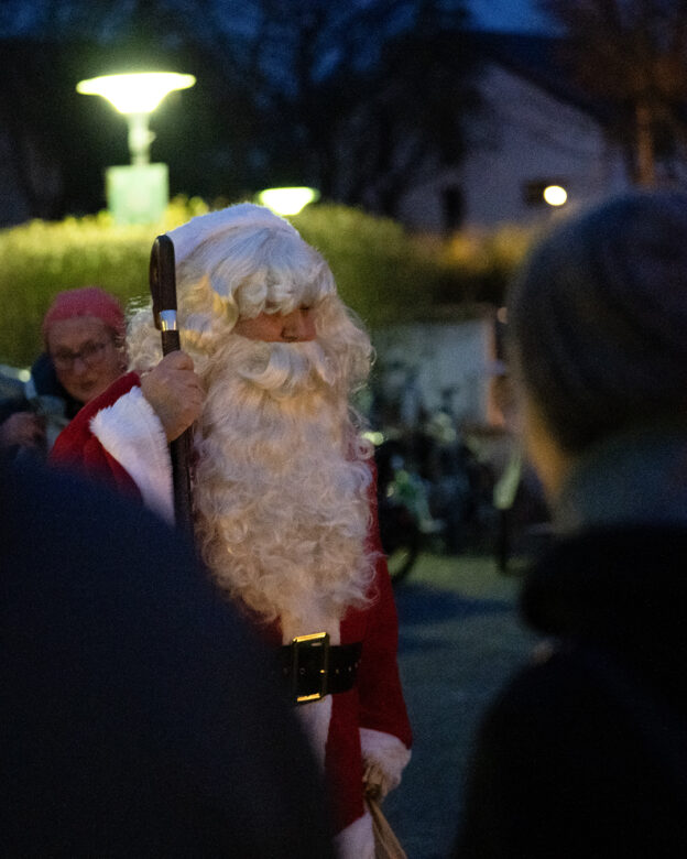 Eine als Weihnachtsmann verkleidete Person hält einen Stab im Freien am Abend, mit unscharfen Menschen und Straßenlaternen im Hintergrund.