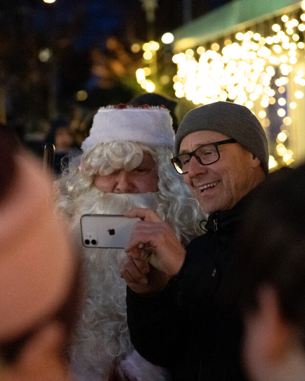 Ein Mann mit Brille und grauer Mütze macht ein Selfie mit einem als Weihnachtsmann verkleideten Mann vor einer nächtlichen Lichterkette.
