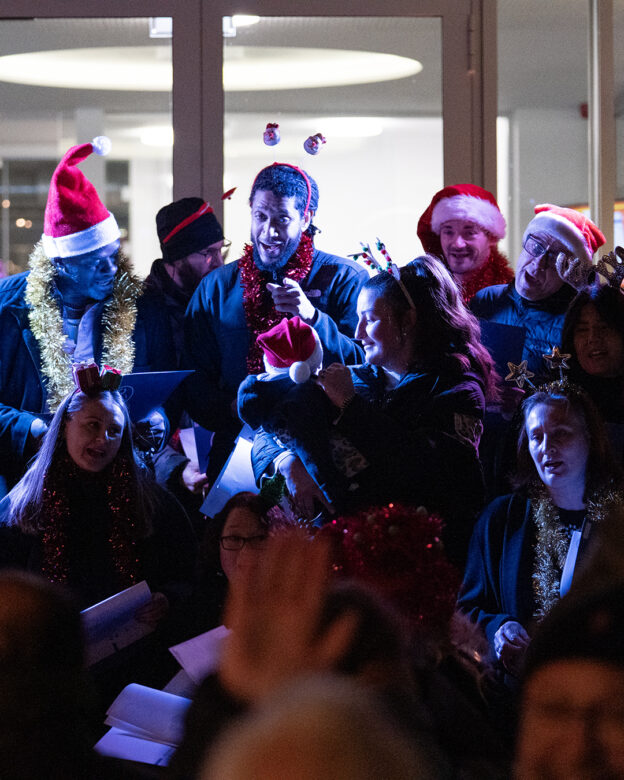 Eine Gruppe von Menschen mit Weihnachtsmannmützen und weihnachtlichen Accessoires singt gemeinsam in einem Haus und hält dabei Liedblätter in der Hand.