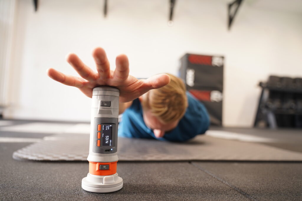 ASC Performance_4 Eine Person in Trainingskleidung greift nach einem Reaktionstrainingsgerät auf dem Boden eines Fitnessstudios, um ihre Leistung zu verbessern, während im Hintergrund Trainingsmatten und Geräte zu sehen sind.