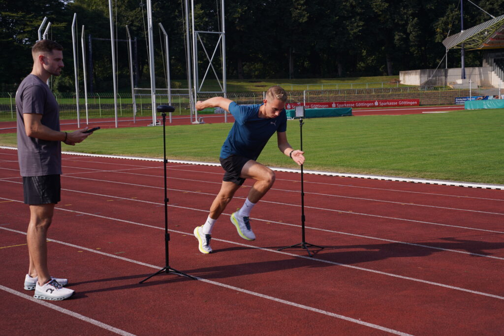ASC Performance_6 Ein Mann sprintet auf einer Bahn von der Startlinie los und zeigt seine Leistung, während ein anderer Mann in der Nähe steht und ein Gerät hält, mit dem er möglicherweise seine Zeit misst.