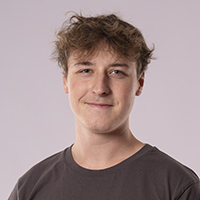 Junger Mann mit kurzen, zerzausten braunen Haaren, der ein dunkles Fitness-T-Shirt trägt, steht vor einem schlichten hellen Hintergrund und schaut mit neutralem Blick in die Kamera.