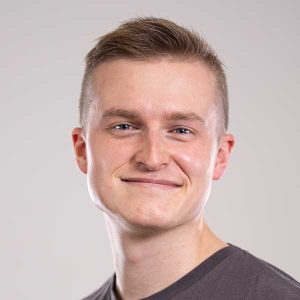 Ein junger Erwachsener mit kurzen, hellbraunen Haaren und heller Haut lächelt in die Kamera. Er trägt ein graues T-Shirt, das vor einem schlichten, hellen Hintergrund dezent seine gute körperliche Verfassung unterstreicht.