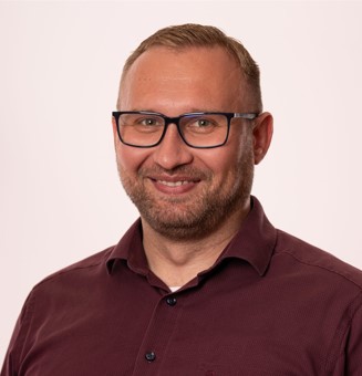 Ein Mann mit kurzen blonden Haaren, Brille und einem gepflegten Bart lächelt in die Kamera. Er trägt ein weinrotes Hemd mit Knöpfen und steht vor einem schlichten, hellen Hintergrund – ein freundliches und professionelles Mitglied des Beratungsteams.