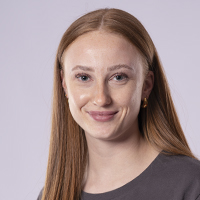 Eine Frau mit langen, glatten, hellbraunen Haaren, die ein graues Fitness-Top trägt, blickt in die Kamera und lächelt leicht. Der Hintergrund ist einfarbig hellviolett.