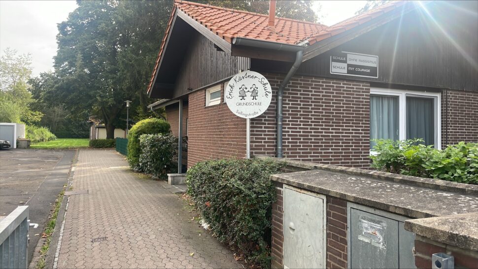 Neben einem gepflasterten Gehweg und einer Grünanlage steht ein Backsteingebäude mit einem weißen, runden Schild, auf dem „Ev. Kindergarten Grünschule“ steht.