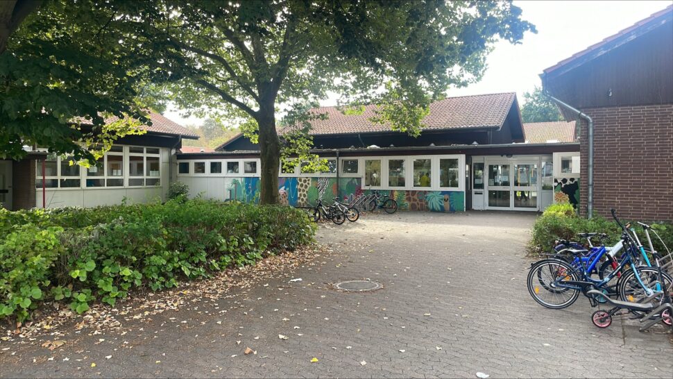 Vor einem Gebäude mit großen Fenstern und blättrigen Bäumen, die einen schattigen Innenhof bilden, stehen mehrere Fahrräder.