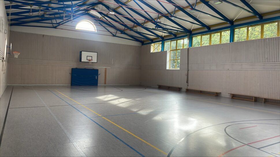 Eine leere Sporthalle mit Basketballkörben, Bänken entlang der Wand und Sonnenlicht, das durch große Fenster und ein Oberlicht hereinströmt. Blaue Balken ziehen sich über die Decke.