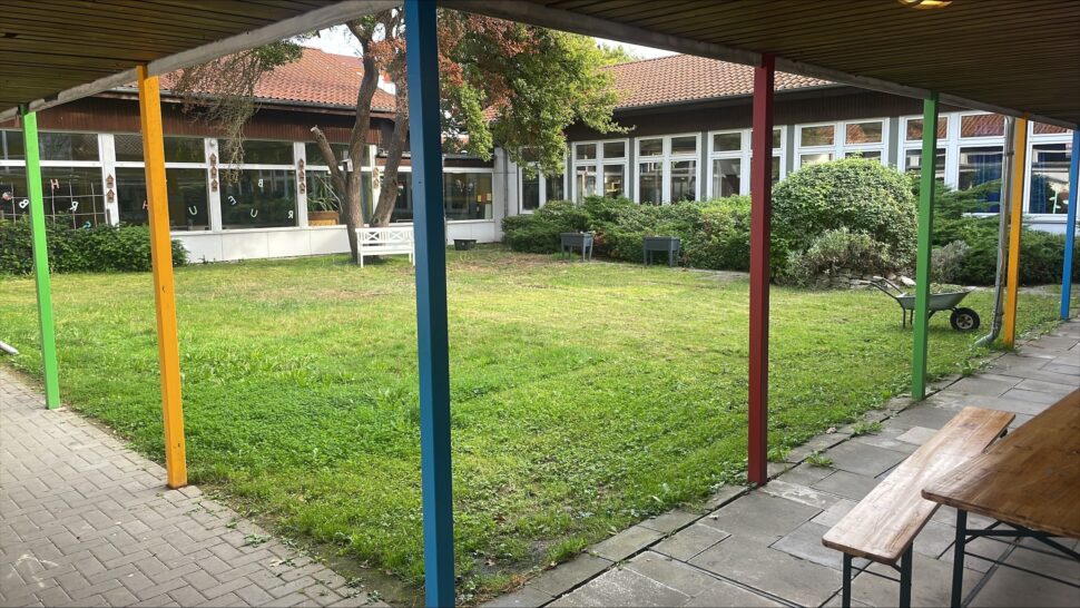 Ein grasbewachsener Innenhof mit bunten Säulen, umgeben von Gebäuden mit großen Fenstern. In der Nähe von grünen Büschen sind eine Schubkarre und Bänke zu sehen.