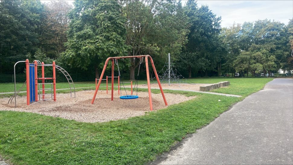 Spielplatz mit Schaukeln, Klettergerüsten und einem Bodenbelag aus Holzspänen, umgeben von Rasen und Bäumen, rechts neben einem gepflasterten Weg.